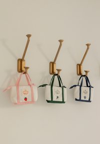 Trois petits sacs en toile blanche avec des poignées et des patchs colorés sont suspendus à des crochets muraux en laiton sur un mur beige.