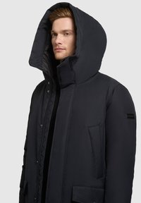 Czarna pikowana kurtka z wysokim kołnierzem i oversize'owym kapturem. Posiada boczne kieszenie oraz naszywkę z logo na rękawie. Gładka struktura.