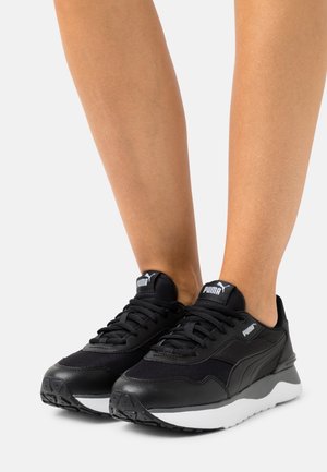 Sneakers nere Puma con suole bianche indossate su gambe nude, viste dalla parte frontale e leggermente di lato su uno sfondo grigio.