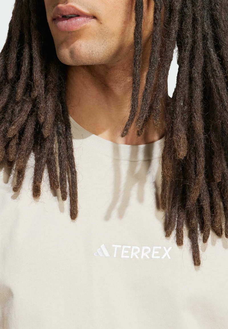 Camiseta de algodón color beige claro con cuello acanalado, que presenta un logo "TERREX" en blanco y un símbolo de adidas bordado en el pecho.
