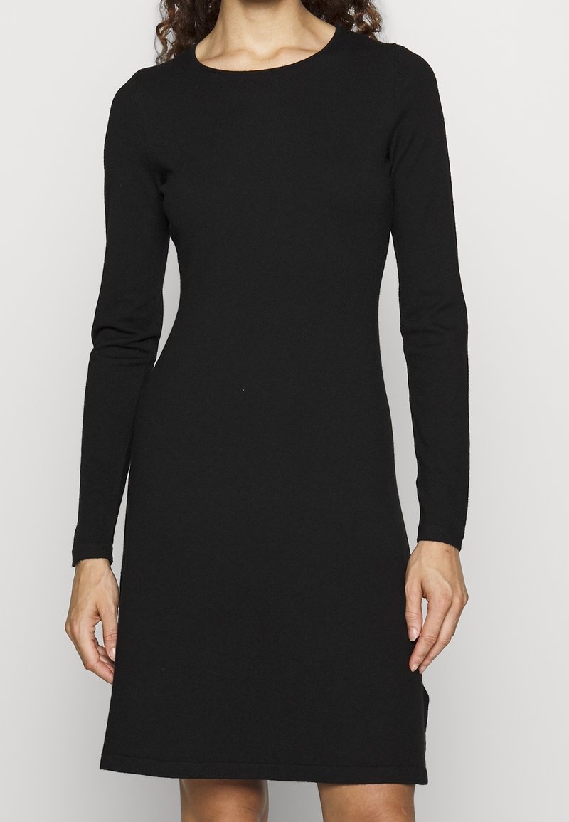 Femme portant une robe noire ajustée, à manches longues, longueur genou, sur un fond clair uni.