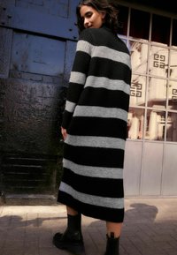 Lange Strickjacke in Schwarz- und Graustripes; gerippte Textur; knielang mit hohem Kragen; kombiniert mit schwarzen klobigen Stiefeln.