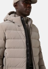 Beige puffer jacket with a hood, featuring horizontal quilted sections, a black zipper, and logo detailing, paired with a gray turtleneck.