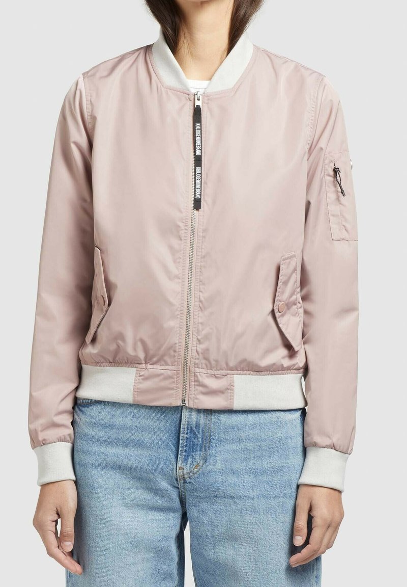 Veste bomber rose clair avec poignets et col côtelés blancs, fermeture éclair à l'avant, poches latérales à rabat, portée avec un jean bleu sur une personne debout.