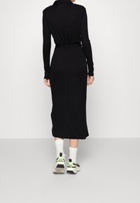 Robe noire à manches longues avec col et taille cintrée, présentant une silhouette ajustée et une longueur mi-mollet. Portée avec des chaussettes blanches et des chaussures de sport.