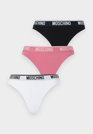 Žena stojaca bosá, oblečená v bledoružovom bralete a čiernom spodnom prádle Moschino s logom na páse na jednofarebnom bielom pozadí.