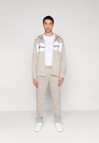 Μπεζ φούτερ με λευκό πάνελ που φέρει το κείμενο "Champion", συνδυασμένο με αντίστοιχο μπεζ παντελόνι φόρμας και λευκά αθλητικά παπούτσια. Απλός, καθημερινός σχεδιασμός.