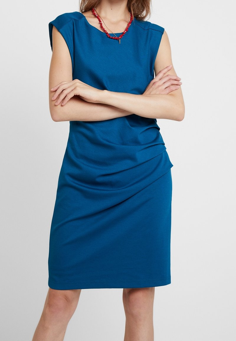 Femme les bras croisés portant une robe bleue sans manches longueur genou et un collier de perles rouges sur un fond clair uni.
