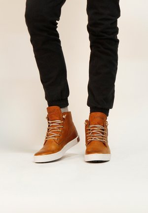 Zapatillas altas de cuero marrón con cordones beige, suelas de goma blancas y acentos texturizados. Llevadas con pantalones negros ajustados.
