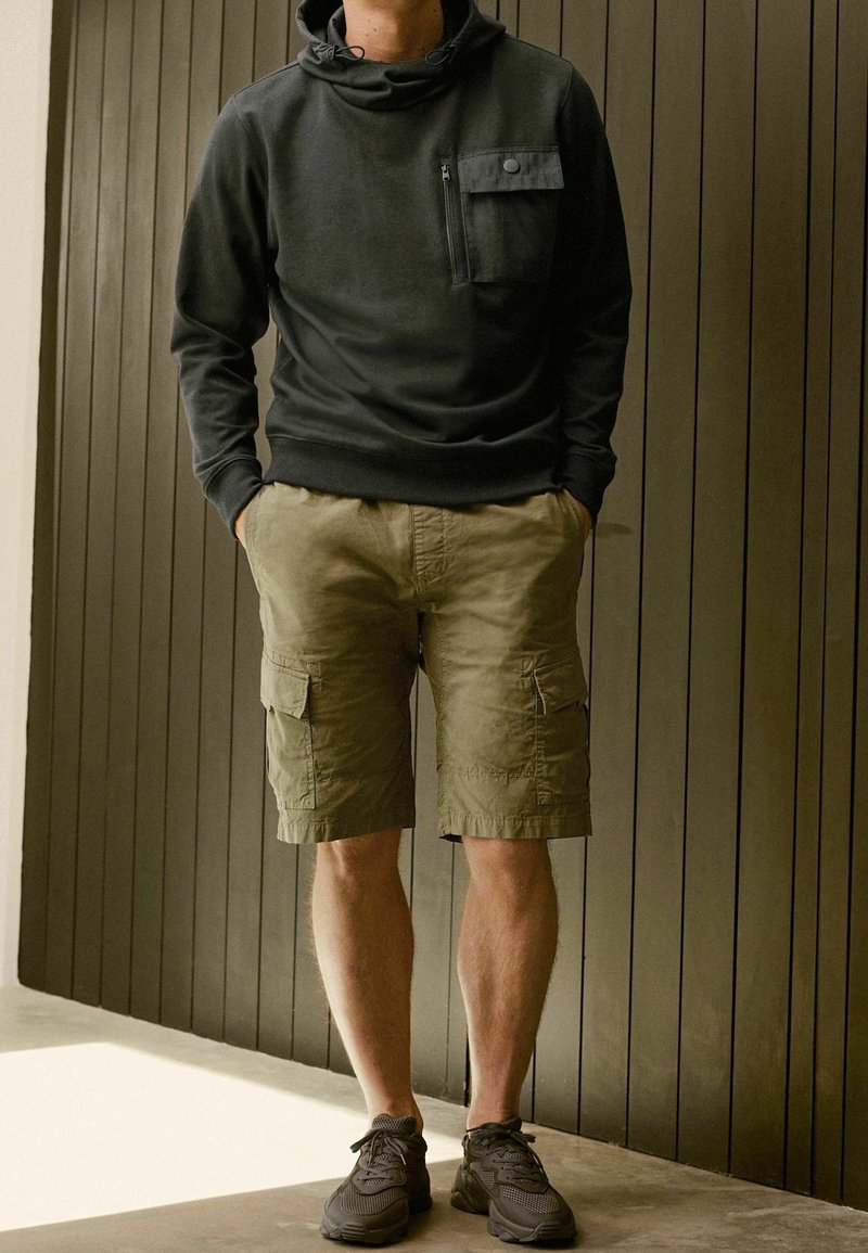 Personne portant un sweat à capuche foncé, un short cargo kaki et des baskets foncées, debout à l'intérieur, devant un mur en panneaux de bois verticaux.