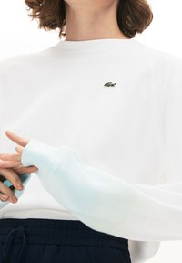 Felpa bianca a maniche lunghe con finitura testurizzata e polsini elasticizzati. Presenta un piccolo logo verde Lacoste sul petto.