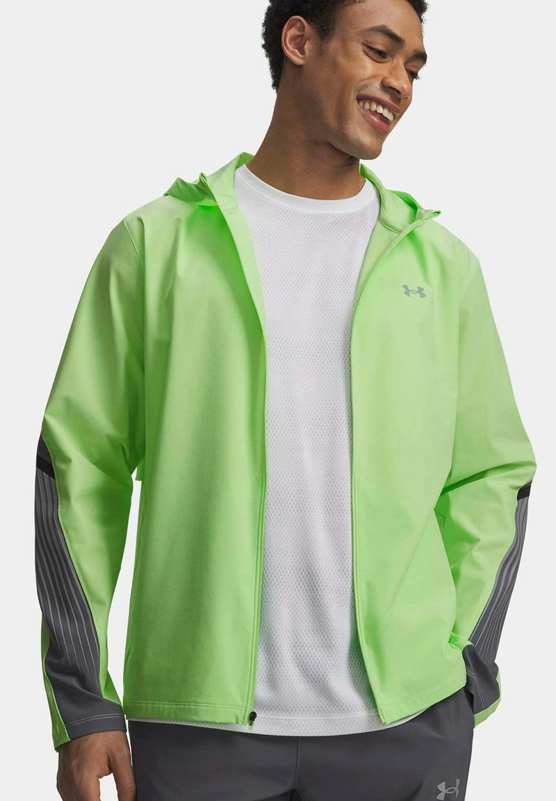 Uomo sorridente che indossa una giacca con cappuccio verde chiaro con maniche a righe grigie, maglietta bianca a rete e pantaloni sportivi scuri su sfondo semplice.