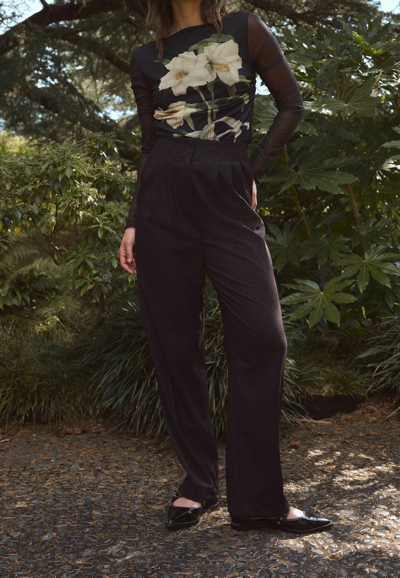 Femme portant un haut noir transparent à manches longues avec un grand imprimé fleur blanche, un pantalon noir taille haute à jambes larges, et des chaussures noires à l'extérieur.