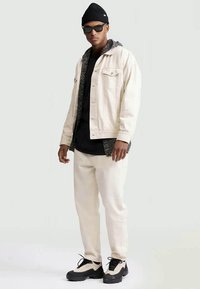 Veste en denim beige avec un hoodie noir en dessous, pantalon blanc et sneakers noirs avec des accents beiges. Le modèle porte un bonnet noir et des lunettes de soleil.