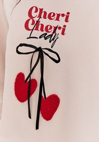 Světle růžová mikina s nápisem "Cheri Cheri Lady" v červeném a černém písmu. Obsahuje černou stuhu a dvě texturované červené srdce připevněná níže.