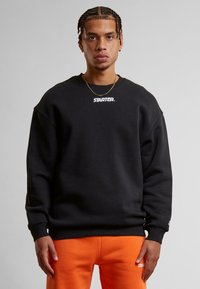 Sweatshirt noir avec épaules tombantes et poignets côtelés, arborant un logo blanc "STARTER." sur la poitrine, associé à un pantalon de jogging orange.