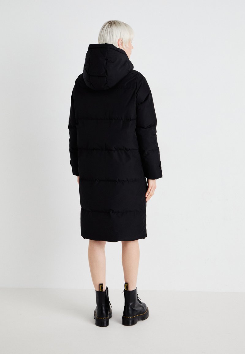Schwarzer Daunenmantel mit Kapuze, mit gestepptem Design und langen Ärmeln, der knapp oberhalb des Knies endet. Trägt man über schwarzen Stiefeletten.