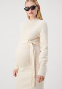 Abito premaman a maglia beige con texture a coste e un dettaglio di vita legata. Le maniche lunghe e lo scollo tondo arricchiscono il design aderente.