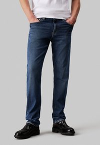 Jeans de denim azul con corte recto, que cuentan con un diseño estándar de cinco bolsillos y costuras visibles. Llevados con zapatos de cuero negros.