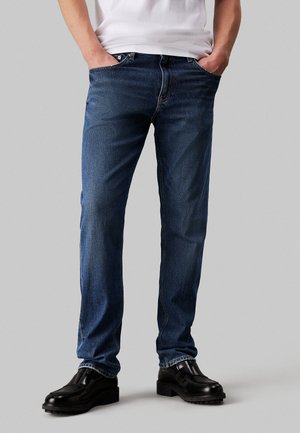 Jeans in denim blu con taglio dritto, caratterizzati da un design a cinque tasche standard e cuciture visibili. Indossati con scarpe in pelle nere.