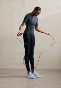 Schwarzes eng anliegendes Kurzarmoberteil, schwarze Leggings und hellblaue Sportschuhe. Die Person hält ein Springseil, vor einem glatten, matten Hintergrund.