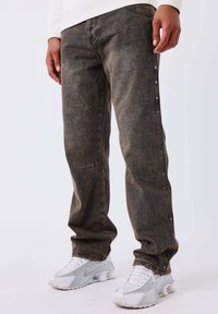 Hombre con jeans rectos de color gris oscuro con botones laterales y zapatillas blancas, de pie contra un fondo claro liso.