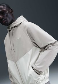 Hombre con chaqueta cortavientos Nike gris claro con capucha, manos en los bolsillos, frente a un fondo gris oscuro.