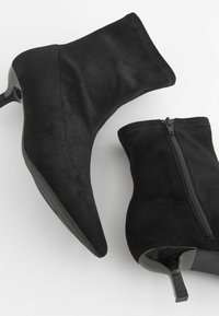 Czarne zamszowe botki za kostkę z ostrym noskiem, smukłym trójkątnym obcasem i zapięciem na zamek. Gładka tekstura z matowymi i błyszczącymi akcentami.