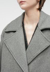 Pilkos vilnonės palto su plačiu įpjautu apykakle ir tekstūruotu paviršiumi, pasižyminčio giliu V formos iškirpimu ir struktūrizuotu, pritaikytu siluetu.