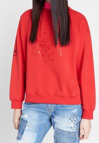 Rode sweatshirt van zacht materiaal, met een verhoogd geometrisch patroon en een hoge halslijn. Gekocht met versleten blauwe jeans.