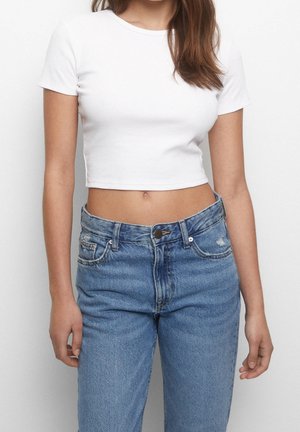 Joven con cabello largo que lleva una camiseta blanca corta de manga corta y vaqueros de mezclilla azul de cintura alta, de pie contra un fondo liso.