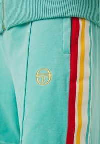 Pantalon de survêtement turquoise avec poches latérales et bande tricotée rayée rouge, blanche, jaune et turquoise sur la couture extérieure.