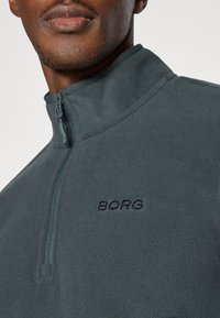 Pullover in pile di colore verde scuro con colletto a zip a un quarto. Presenta il testo ricamato "BORG" sul petto. Texture morbida.
