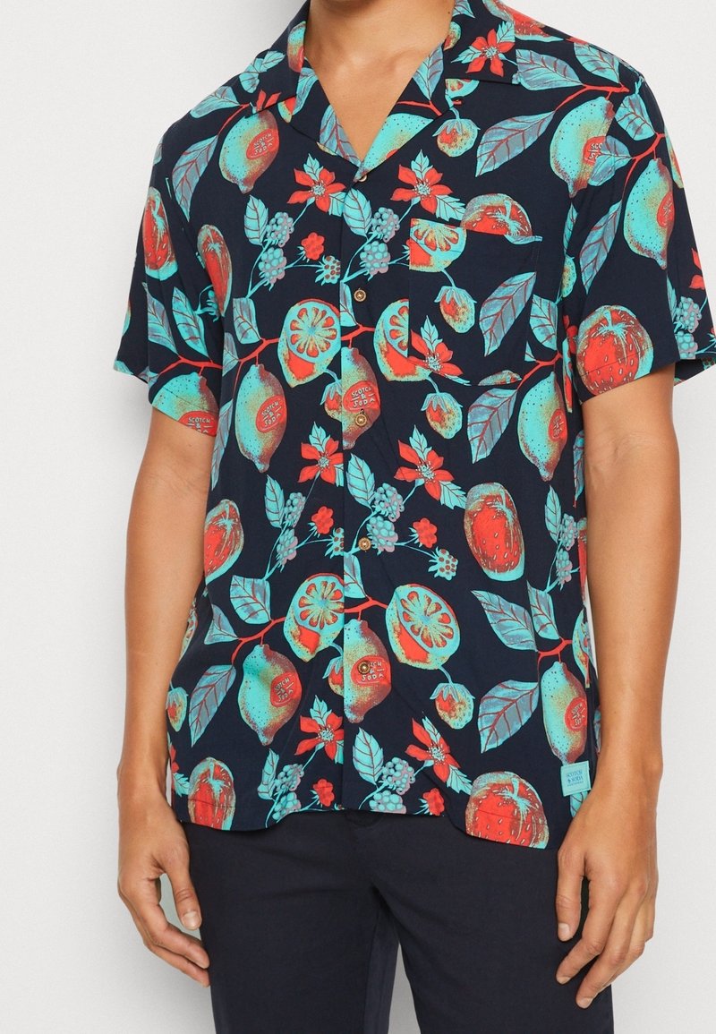 Lyhythihainen, napillinen paita, jonka kankaan väri on tummansininen ja jossa on kirkkaanvärisiä hedelmä- ja kukkakuoseja punaisessa ja turkoosissa, sileä materiaali, rento istuvuus.