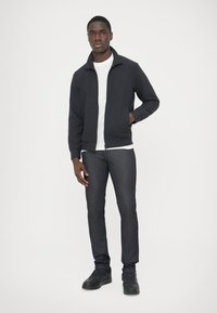 Σκούρο καπιτονέ μπουφάν με φερμουάρ πάνω από ένα λευκό μπλουζάκι, σε συνδυασμό με σκούρο τζιν και μαύρα παπούτσια. Το μοντέλο στέκεται σε ουδέτερο φόντο.