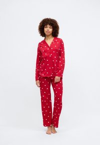 Κόκκινο σετ πιτζάμας με μακριά μανίκια και παντελόνι, διακοσμημένο με λευκό μοτίβο καρδιάς. Κατασκευασμένο από μαλακό ύφασμα με άνετη εφαρμογή.