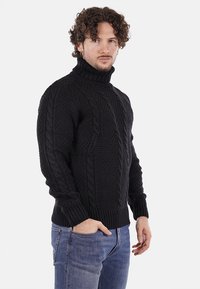 Maglione a collo alto intrecciato di colore nero, realizzato in filato testurizzato, con orlo e polsini a coste, indossato con jeans blu chiaro.