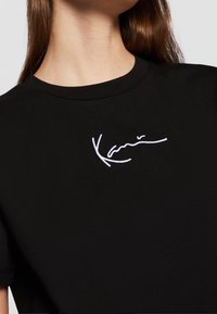 T-shirt noir à manches courtes en tissu doux, avec une signature brodée blanche sur la poitrine, un col rond et une coupe décontractée.