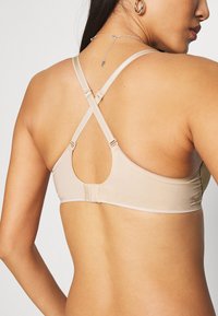 Beige sportbeha met een gladde textuur, kruislingse banden op de rug en een haaksluiting. Voorzien van een uitsnede-ontwerp op de rug.