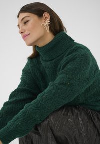 Zaļš adīts turtlenecks džemperis ar teksturētu rakstu, apvienots ar melnu plīsušo svārku; modele valkā lielu, lentveida, pērļu līdzīgu auskaru.