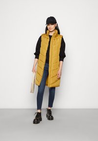 Colete puffer dourado com quilt vertical, gola alta e fecho na frente. Combinado com jeans escuros e botas pretas de laço. Boné preto e bolsa pequena.