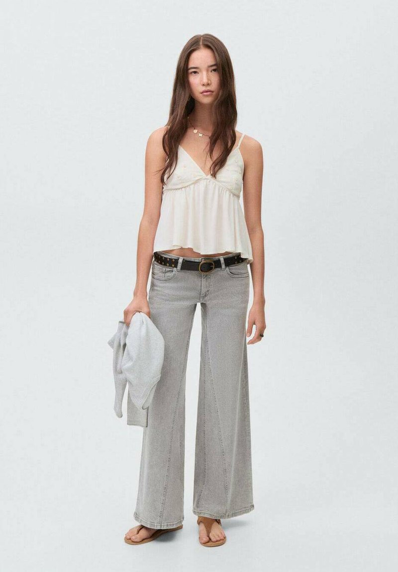 Vit kort camitopp med justerbara axelband, matchad med vida ljusgrå jeans. Modellen håller en grå tröja och har sandaler på sig.