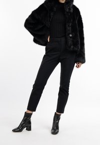 Schwarze Kunstfelljacke mit geripptem schwarzen Oberteil, maßgeschneiderten schwarzen Hosen und schwarzen Absatzstiefeletten. Strukturiertes, warmes und figurbetontes Design.