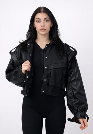 Jonge vrouw met lang zwart haar, gekleed in een zwarte cropped leren jas over een zwart outfit met rits, staand tegen een witte achtergrond.