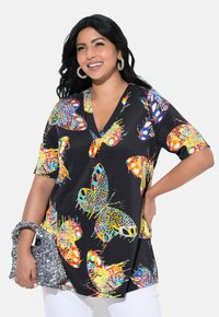 Sort tunika med farverigt sommerfuglemotiv, der har V-udskæring og korte ærmer. Modellen holder en glitrende grå taske, som er parret med hvide bukser.