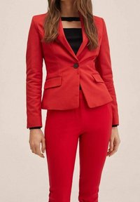 Blazer rouge à coupe ajustée, bouton noir unique, et deux poches avant, associé à un pantalon rouge sur mesure. Texture de tissu lisse.