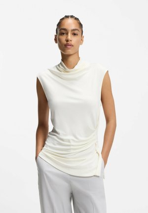 Hihaton kermaasuinen paita, jossa laskostettu pääntie ja sivussa kerätty yksityiskohta, valmistettu sileästä kankaasta. Yhdistetty vaaleanharmaisiin housuihin.