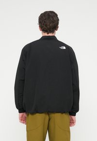 Μαύρο ελαφρύ μπουφάν με όρθιο γιακά, ελαστικά μανίκια και διακριτικό λογότυπο στην πλάτη. Ματ φινίρισμα, φορεμένο πάνω από ελιά παντελόνια.