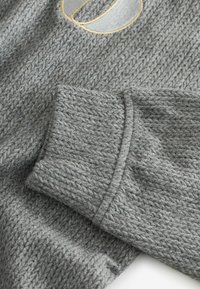 Šedý pletený svetr s detailním texturovaným vzorem, ribbed manžetami a kontrastní vyšívanou nášivkou poblíž výstřihu.