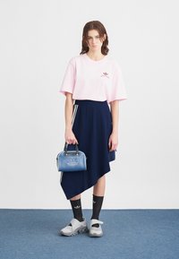 Jauna sieviete, ģērbusies rozā Adidas T-kreklā, tumši zilā asimetriskā svārkā, melnās Adidas zeķēs, pelēkās kedās un tur rokās zilu Adidas rokassomu.